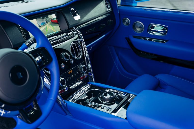 Blue luxury car interior with blue leather seats, steering wheel, and a chrome-accented center console. A rearview camera display is visible on the dashboard screen, showing a parking view.