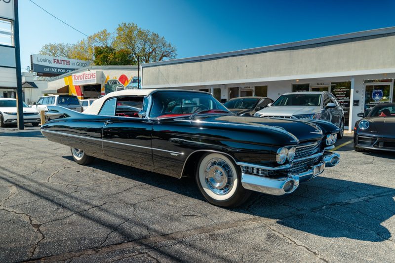 Shiny black 1950s Cadillac convertible with white roof in a parking lot, chrome accents and whitewall tires, with other classic cars nearby.
